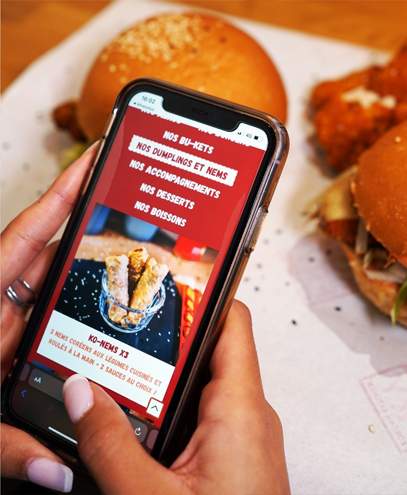 Téléphone affichant le menu d'un restaurant, avec des burgers en arrière-plan, photographié par l'agence de communication Glick Légende :